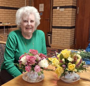 A lady in a green top with two teacup displays of flowers. One with pink and white flowers and the other yellow flowers